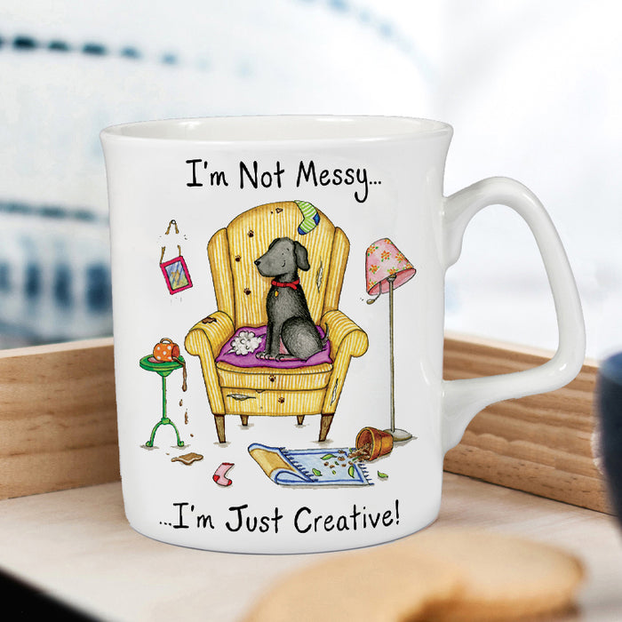 A white ceramic coffee mug with a humorous illustration of a grey dog sitting in a yellow armchair, surrounded by scattered toys and the text 'I'm Not Messy.